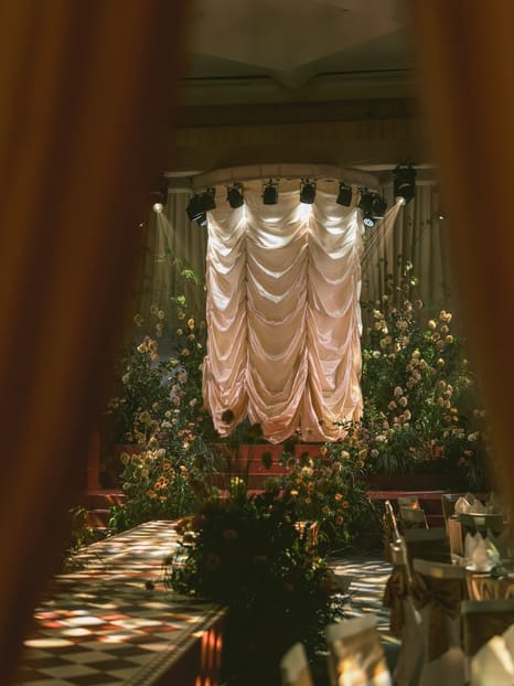 Blush satin ceremony backdrop framed by curtains at a floral reception