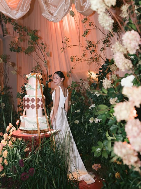 Bride beside a tiered wedding cake in a floral-lit reception setting